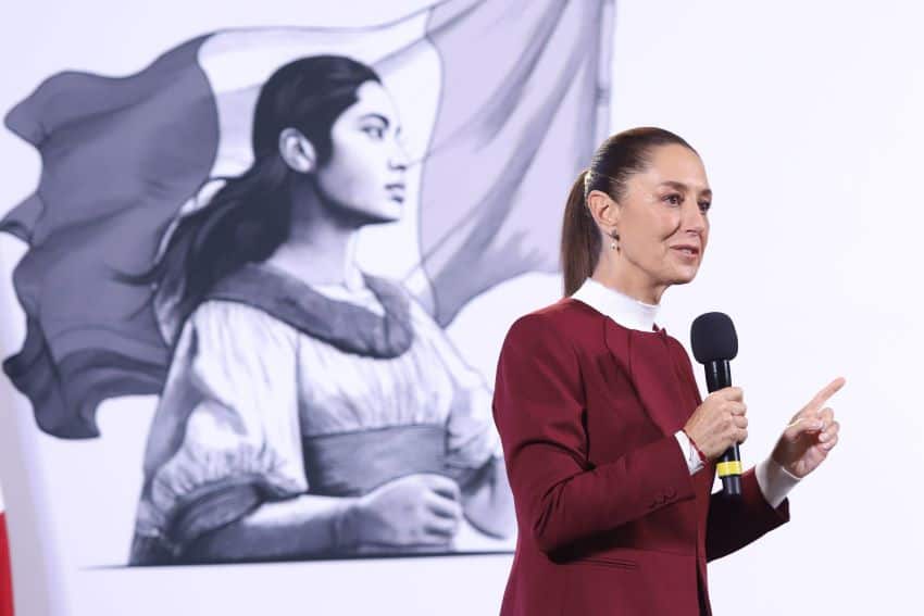 Sheinbaum wants Spain’s apology: the mañanera recapped President Claudia Sheinbaum holding a microphone in front of her administration's logo of a Indigenous Mexican woman in front of the Mexican flag.