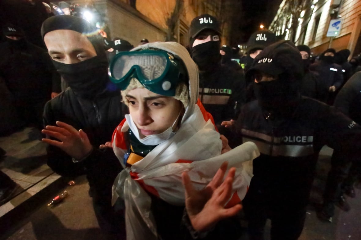 A protester wearing protective goggles and draped in a Georgian flag is pushed forward by a crowd of masked police. 