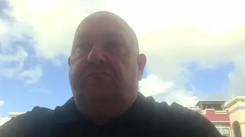 A zoom screenshot of Billy George - he is bald and is wearing a black polo shirt. Behind him is a blue sky with white clouds.