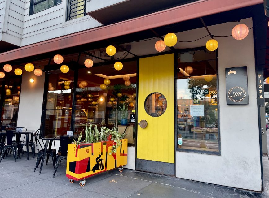 A restaurant entrance with a yellow door, round window, and hanging lights. Outdoor seating is visible, and a planter cart with greenery is in front.
