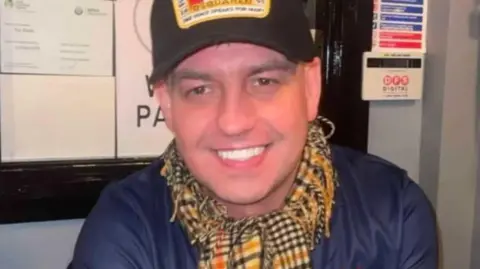 Family handout John Hardy is smiling into the camera. He's wearing a black cap, with a design on it, and a navy top and a checked scarf.