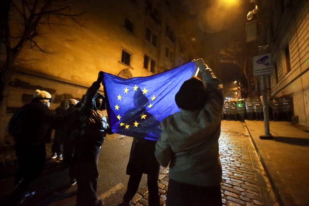 A small group in an alley hold up a blue flag featuring a circle of yellow stars as they stand in front of a group of police with riot shields at the other end of the street.  