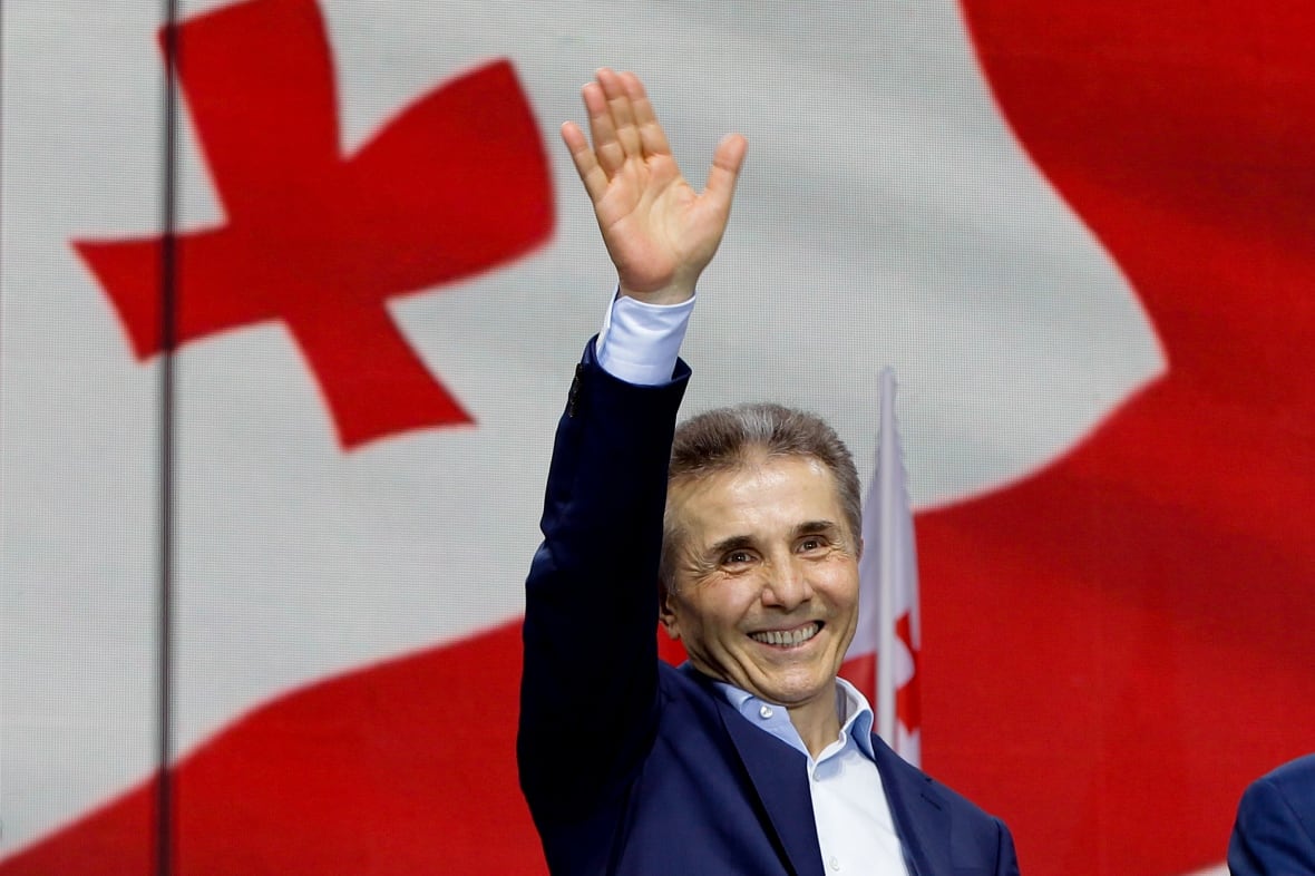 A man in a suit and short brown hair waves while standing in front of a Georgian flag. 