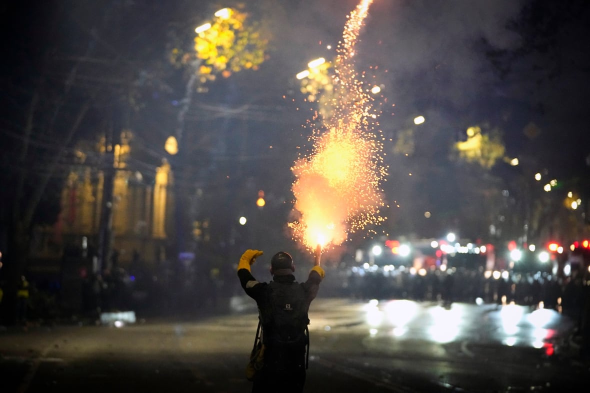 A person wearing dark clothing and bright yellow gloves stands with their back to the camera holding a firework and facing off against a line of law enforcement with shields in the distance. 