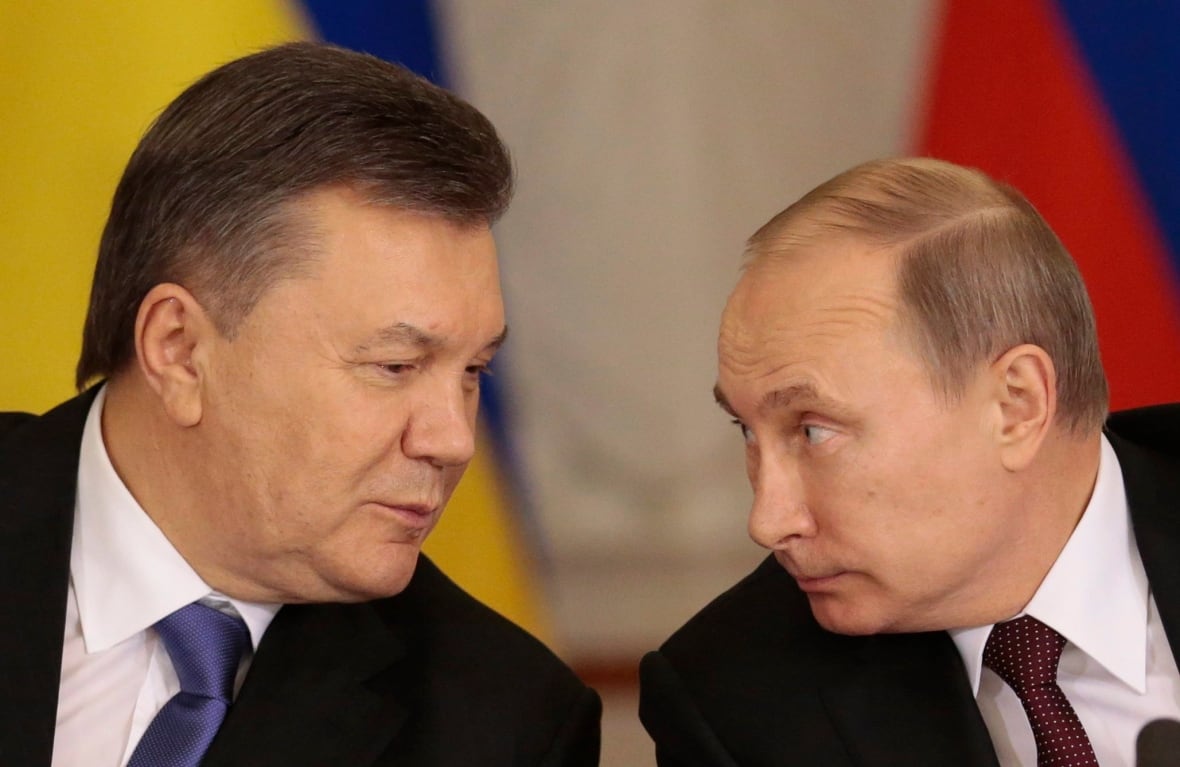 Two men in suits speak in front of flags. 