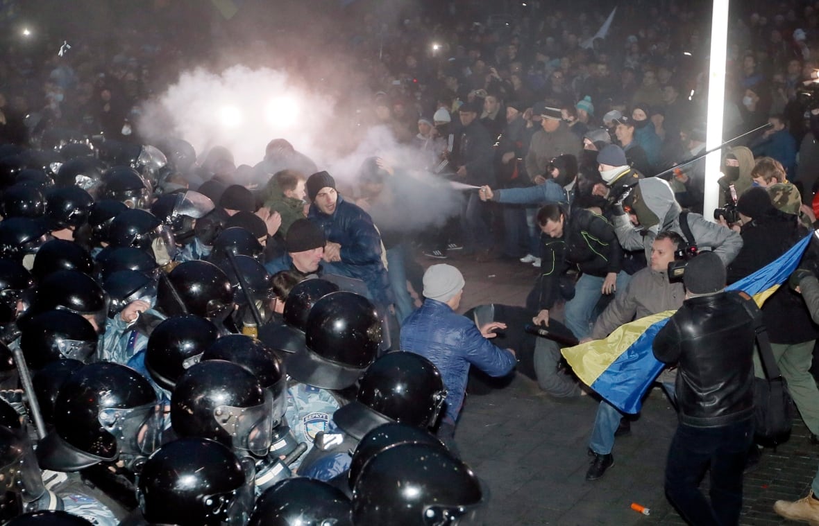Crowds of people square off against each other in an outdoor square, spraying teargas and pushing and shoving as police in riot gear look on. 