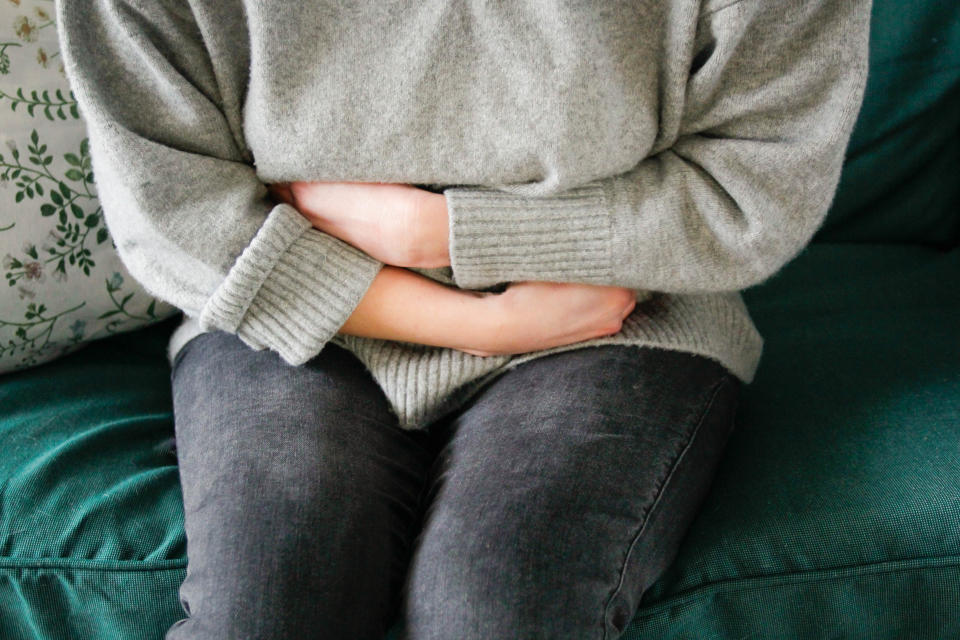 A woman with cramps wraps her arms around her abdomen as she sits on a couch.