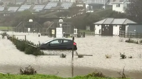 Keith Smith A black car in brown flood water up to the wheel arches. There is no one in the vehicle. A flooded play park can be seen in the background.