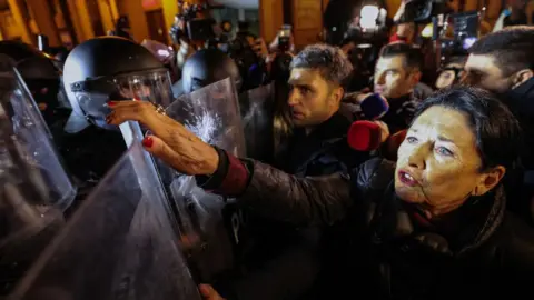 Getty Images Georgia's President Salome Zourabishvili, a woman with black hair, stands directly in front of riot police, reaching up over a shield towards a masked officer.