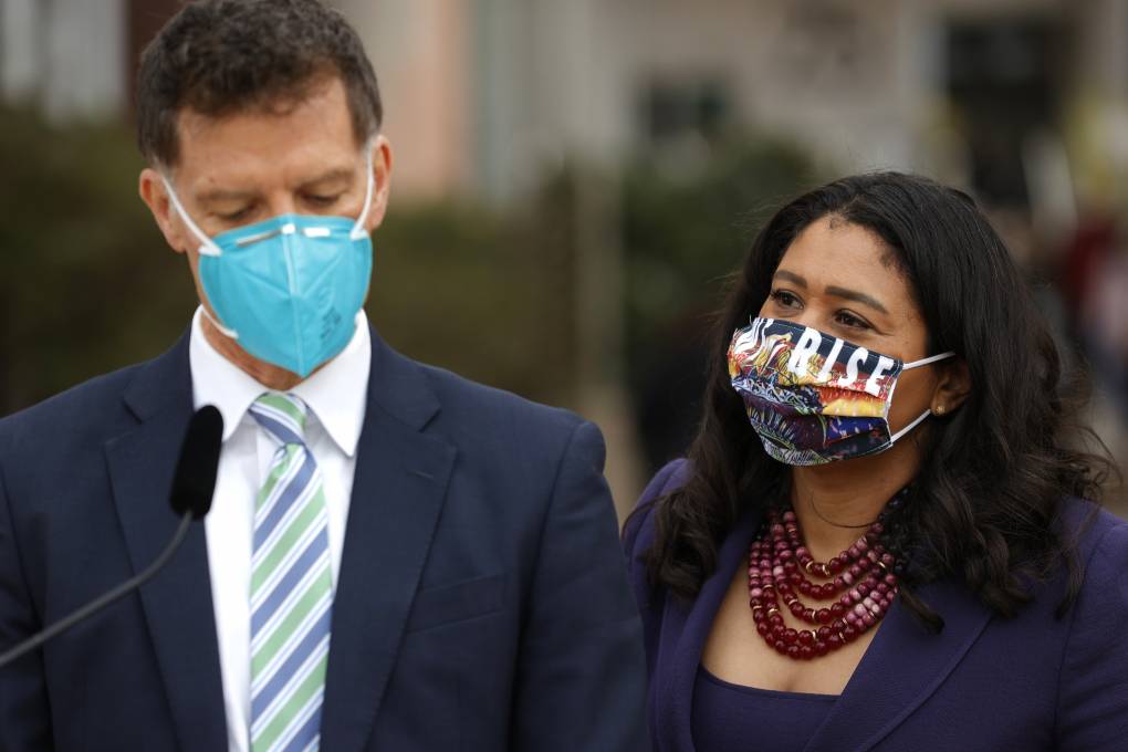 San Francisco Director of Public Health Dr. Grant Colfax Resigns A man and woman stand next to each other. The man is wearing a business suit and light blue mask in front of a microphone and the woman is wearing a navy blue jacket with a red beaded necklace and multicolored mask.