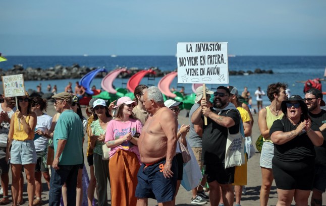 A protester holds a placard reading "Invasion does not come in 'patera', it comes by plane" as thousands march on Las Americas beach during a demonstration to protest against mass tourism, in Arona on the Spanish Canary island of Tenerife, on October 20, 2024. Thousands of flag-waving demonstrators hit the streets across Spain's Canary Islands today to demand changes to the model of mass tourism they say is overwhelming the Atlantic archipelago. Rallying under the slogan "The Canary Islands have a limit", demonstrators waving white, blue and yellow flags of the Canary Islands, marched by tourists sitting in outdoor terraces in Playa de las America before they rallied on the beach chanting "This beach is ours" as tourists sitting on sunbeds under parasol shades looked on. (Photo by DESIREE MARTIN / AFP) (Photo by DESIREE MARTIN/AFP via Getty Images)