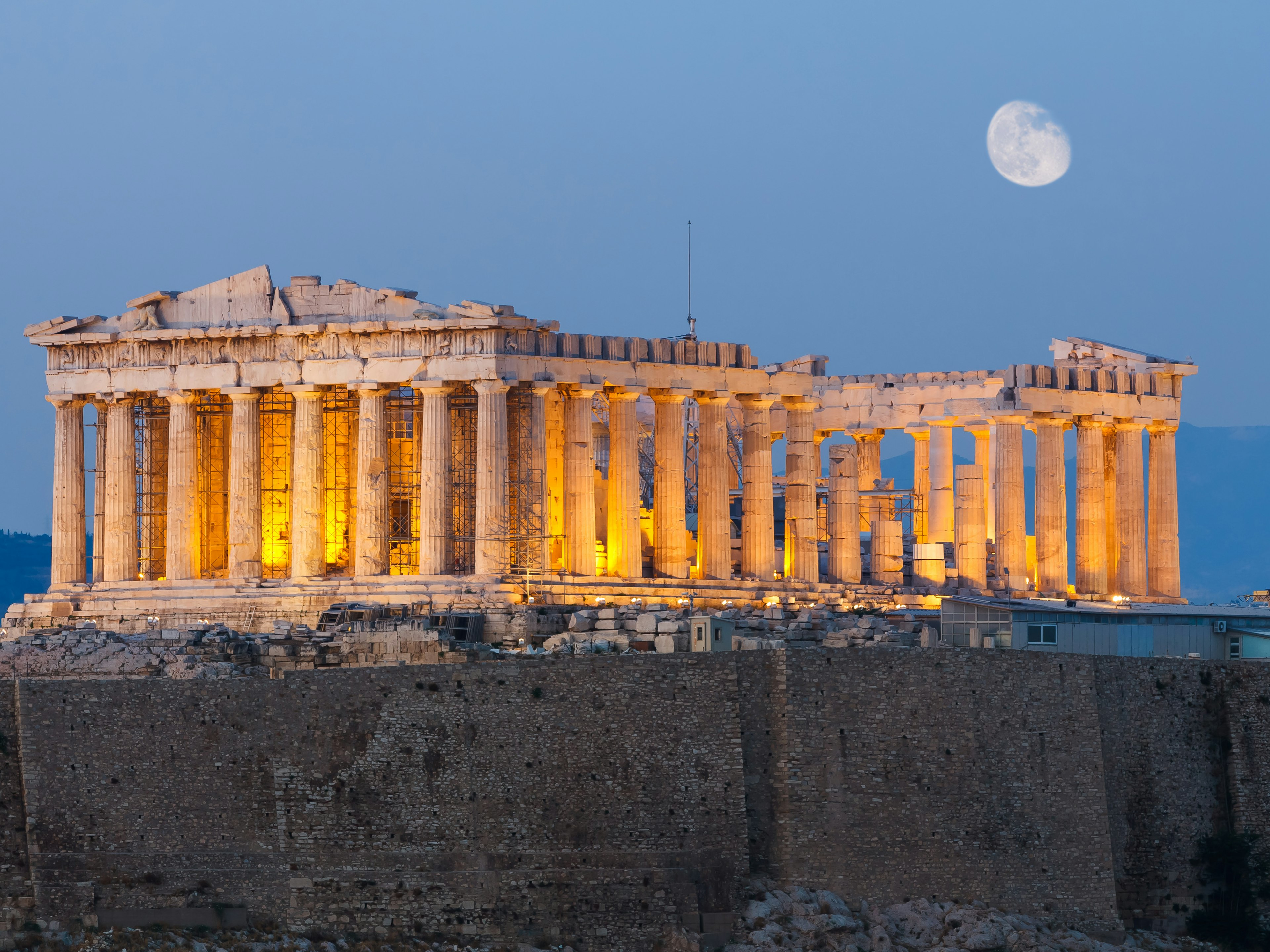 The lit-up structure of the Parthenon at night, with the moon in the background. 