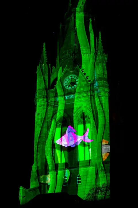 Ian Georgeson Clock tower lit up green, with a pink fish 'swimming' across it.