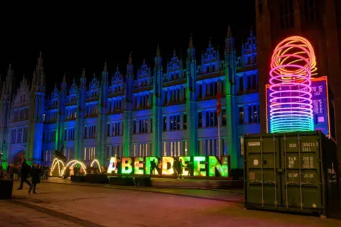Ian Georgeson Impressive granite Marischal College building lit up blue, with the word 'Aberdeen' in block capitals also lit up, and a bright slinky-style object.