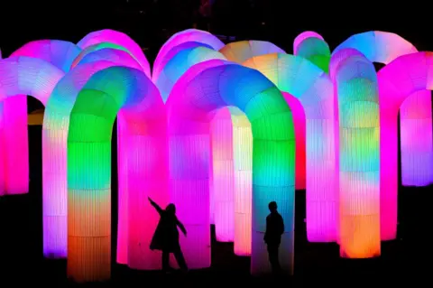 Ian Georgeson Silhouettes of two people amid brightly-coloured tubes - pinks and blues and oranges and greens.