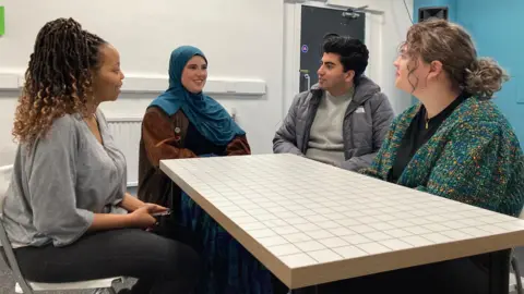 BBC The women and a man sit around a table talking.