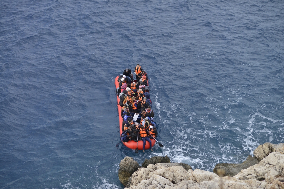 A crowded inflatable boat carrying people in life vests approaches a rocky shoreline on open water.