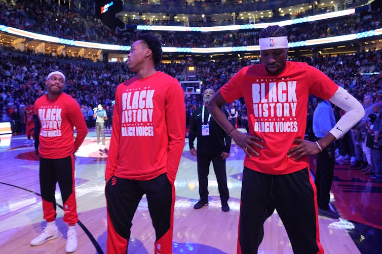 Two black men in red long sleeve shirts look at each other while standing on a basketball court in front of a full stadium