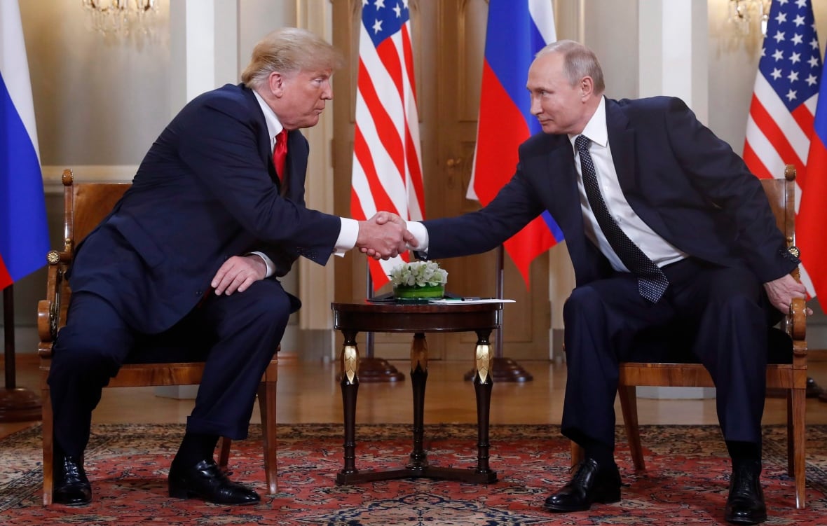 Two men shake hands across a table, with flags in the background.