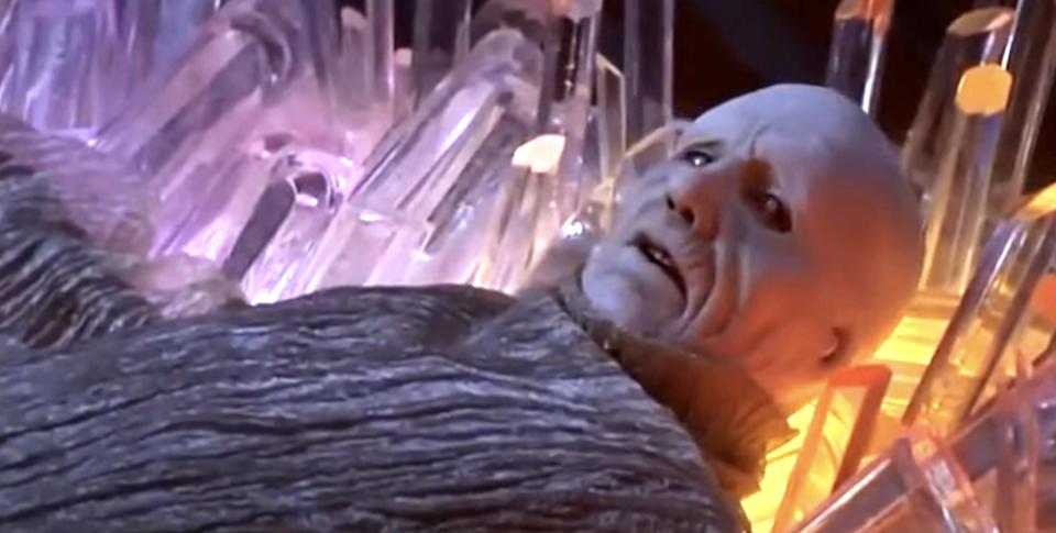 A character with a bald head and wrinkled face is lying in a bed of crystal-like structures, appearing surprised or concerned