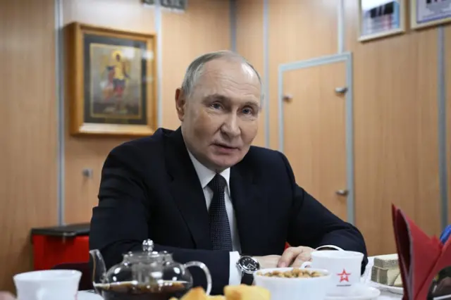 Putin sitting at a table, which has on it a teapot of brown liquid, teacups on saucers and a bowl of snacks. He has his arms sat on the table in front of him and looks over the camera. The room has wooden walls and a few framed pictures