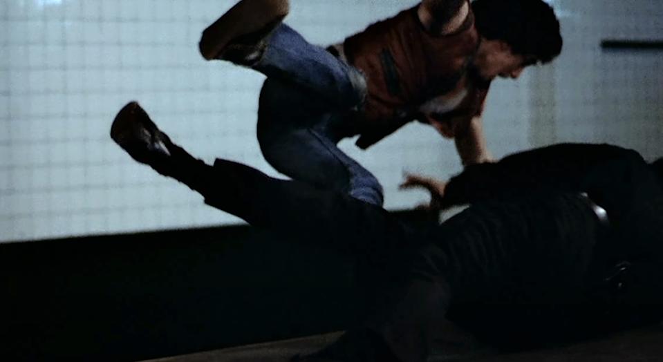 A person in jeans jumps towards another on the ground in a dramatic, action-packed scene, suggesting a fight