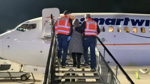 An Garda Síochána Two men in orange high-viz vests with back-up team written on the back lead a woman in a grey coat and dark trousers up the steps of a plane