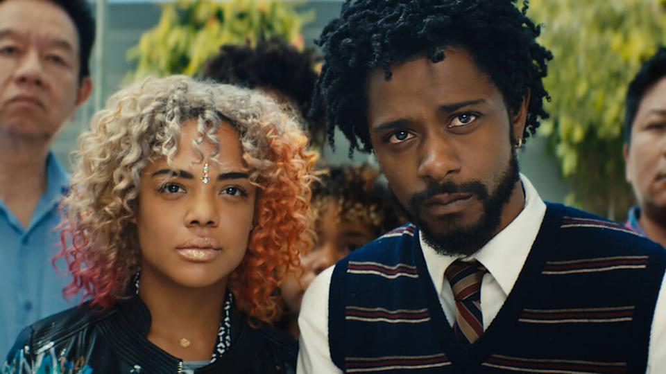 Two individuals stand close together in a crowd of people. The person on the left has curly blonde and orange hair, a necklace, and a black and blue patterned top. The person on the right has curly black hair, a beard, and wears a dark striped sweater vest over a white shirt and tie. Both individuals have serious expressions. Behind them, blurred figures of other people are visible, and the setting appears to be outdoors with greenery in the background.
