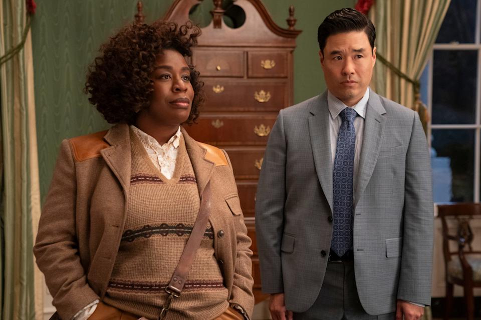 uzo aduba and randall park in the residence. aduba is wearing a beige sweater vest and suite, park is in a grey suit. they're standing in a room with old furniture