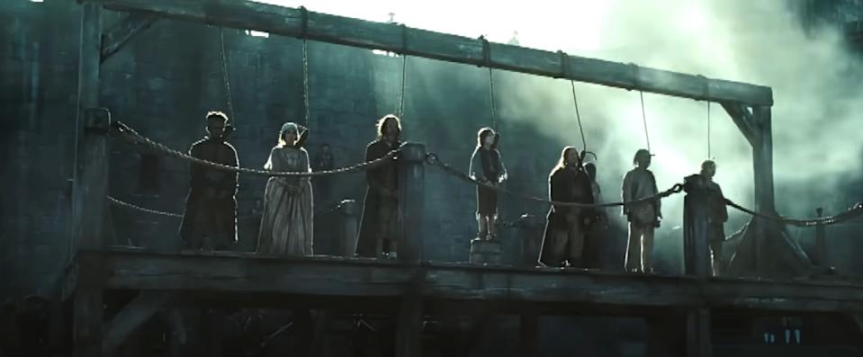A group of people in period clothing stand solemnly on a wooden platform with ropes, appearing as if in a dramatic scene from a historical film