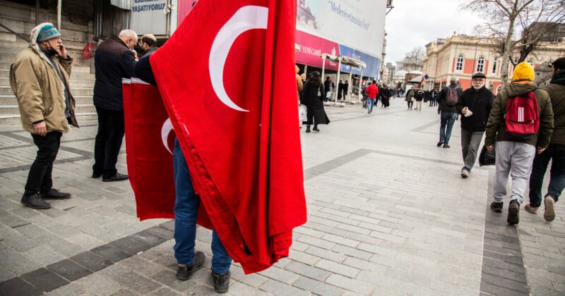 A person stands in a plaza holding several large Turkish flags, obscuring their body. People walk past on the gray cobblestone street, some wearing winter clothing. A building with columns is in the background. The scene feels casual and busy.
