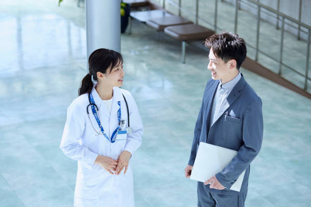 Doctor in white coat with stethoscope conversing with businessman holding laptop in a modern hospital lobby.