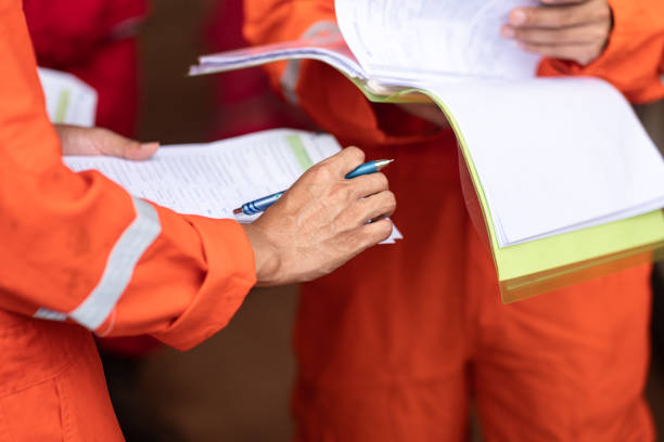 Workers in orange safety uniforms reviewing documents and making notes on a clipboard with pen.
