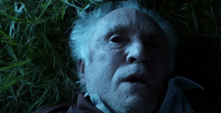 Elderly man lying on grass, looking upwards with a serious expression