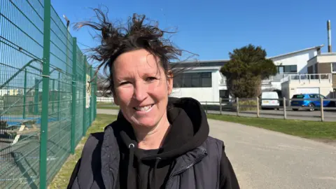 Vicky Barratt is standing outdoors on a sunny day, smiling at the camera. She has dark brown windswept hair and is wearing a black hoodie layered under a dark-coloured vest. In the background is a green metal fence, a paved path, a modern white building with large windows, and several parked cars.