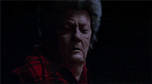 An older character in a plaid shirt closes their eyes dramatically in a dark scene from a TV or movie