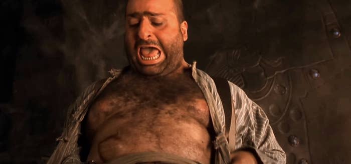 A person in a dramatic movie scene looks down in alarm, wearing an open shirt and suspenders, against a dimly lit background