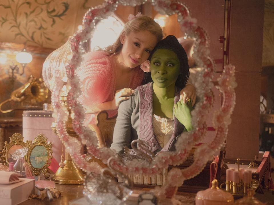 A still from "Wicked" shows Ariana Grande wearing a pink dress and Cynthia Erivo, painted green, wearing a purple robe. They are reflected in a mirror on a table.