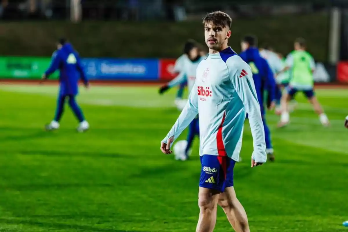 A player in a white and blue training uniform walks across the field while other players practice in the background.