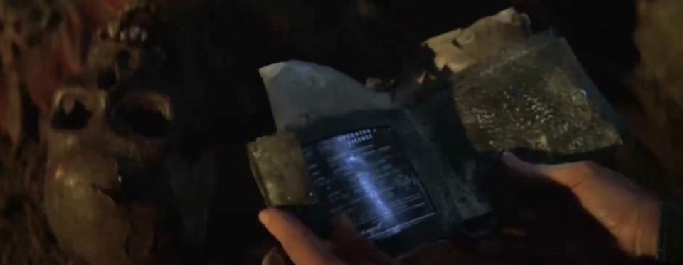 Person holding a futuristic, weathered device in a dark setting, displaying digital information. Skulls are faintly visible in the background