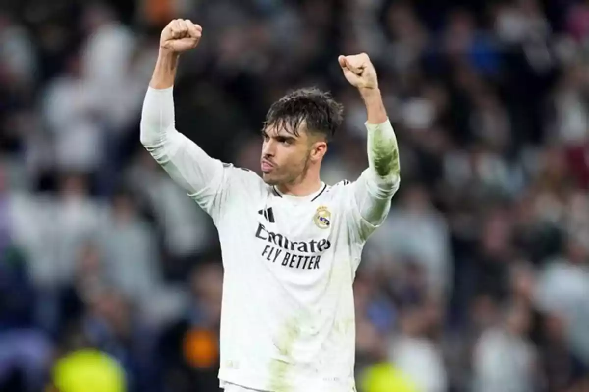 Soccer player in a white uniform celebrating with arms raised in a stadium.