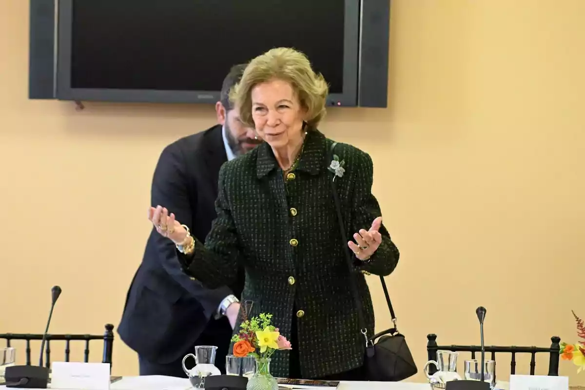 Queen Sofía in a dark coat is standing in front of a table with flowers and glasses, while a man is behind her.