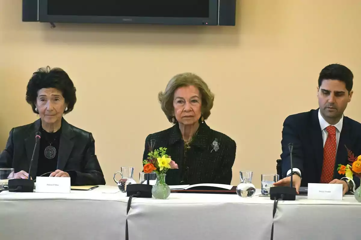 Queen Sofía along with two other people seated at a table with microphones and flowers, participating in a formal event.