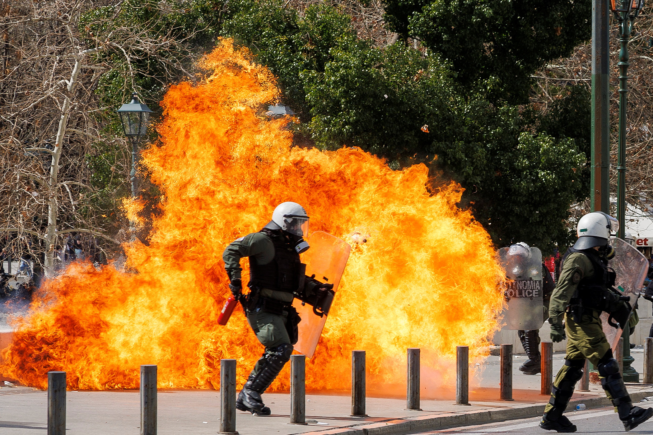 Riot police officers clash with protesters amidst a large fire during a demonstration in Athens.