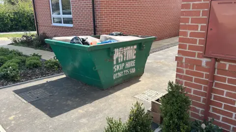 Gemma Allen A green skip on a driveway in front of a house. It is full of rubbish and has the words "Pryme Skip Hire" and a phone number on the side in white bold lettering.