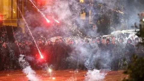 Reuters Fireworks land near a barricade of police, who are in riot gear and holding up shields
