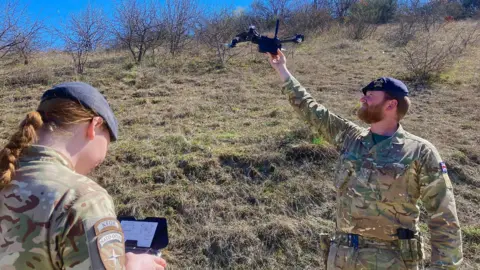 BBC A British soldier in camouflage uniform is preparing to hand launch a reconnaissance drone in Kosovo. Another soldier has the remote control unit. 
