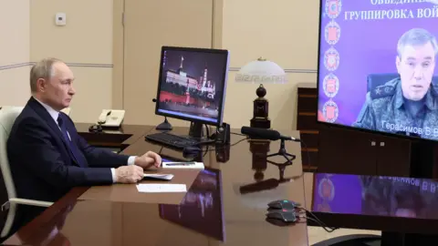 EPA Vladimir Putin on a video call with his army chief in his office, the former sat behind the desk and the latter on a screen.