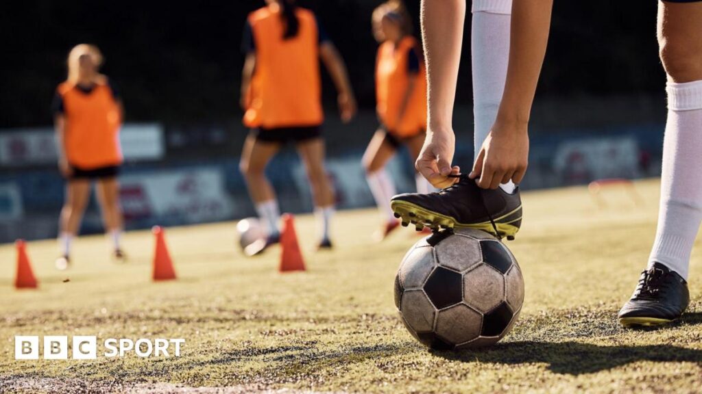 A female footballer bends down to tie her boot, which is resting on the ball while her team-mates train in the background.