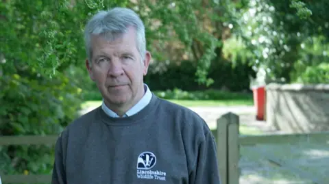 A man wearing a grey jumper with a Lincolnshire Wildlife Trust logo on. He has a white shirt underneath and has short grey hair. He is standing next to a wooden gate with trees in the background.